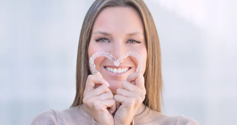 A woman holds two clear aligner trays to form a heart shape and smiles